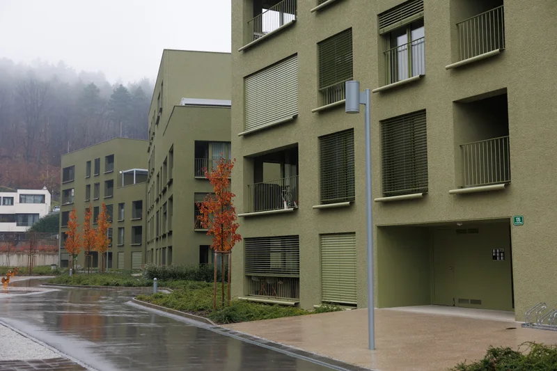 Zelene fasade, lože, drevesa in tlakovana pešpot med stanovanjskimi stavbami.