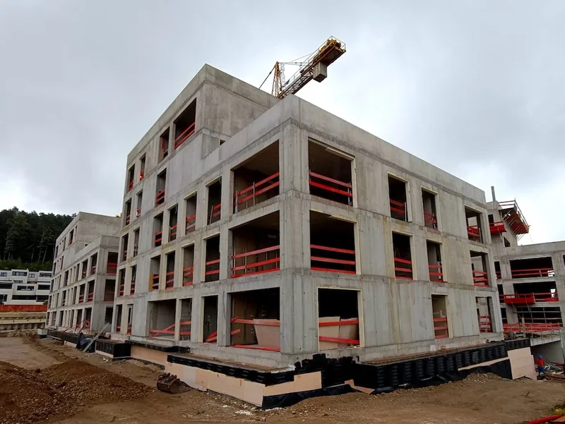 Betonska konstrukcija večstanovanjske stavbe z odprtimi okenskimi odprtinami, rdečimi varovalnimi ograjami in žerjavom.