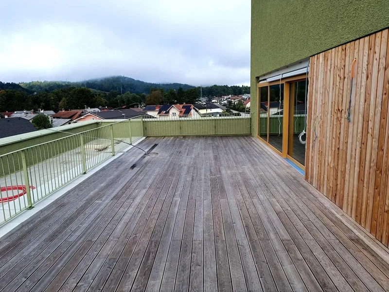 Lesena terasa z zeleno ograjo in navpično macesnovo fasadno oblogo ob zasteklitvi.