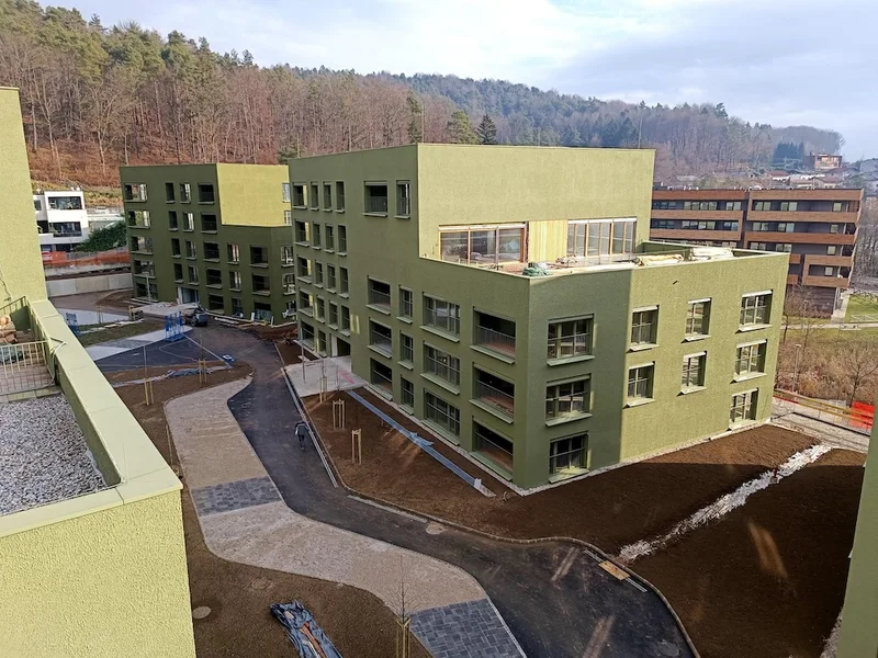 Pogled z višine na zelene stavbe, zavite poti in novo zasajena drevesa; v ozadju je gozdnat hrib.