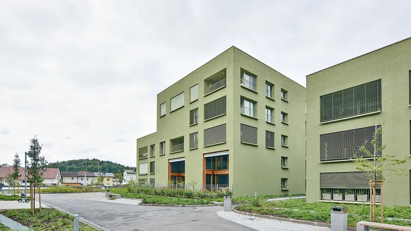 Razvejana pot in zasaditve med zelenimi stanovanjskimi stavbami z ložami in velikimi pritličnimi okni.
